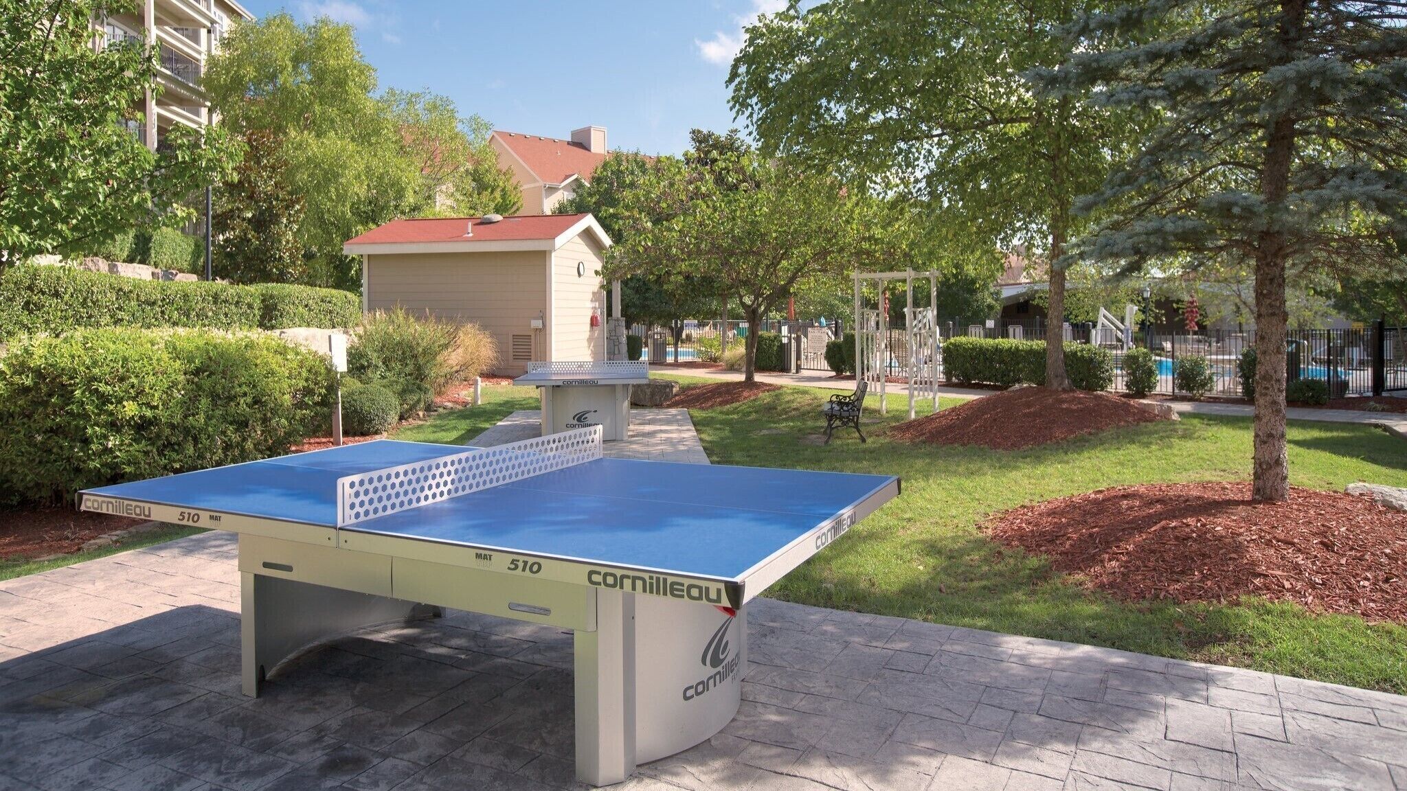 An outdoor table tennis table labeled 'Cornilleau 510 MAT' in a park setting with trees and a building in the background.