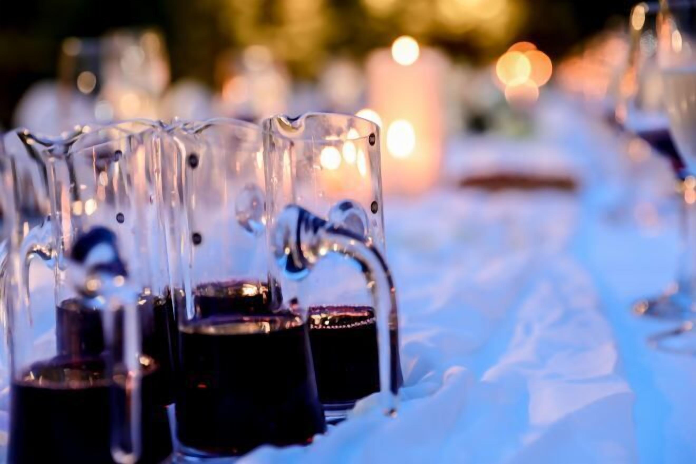 A row of clear glasses filled with a dark liquid, possibly wine, on a table with a blurred background of lights.