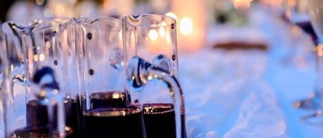 A row of clear glasses filled with a dark liquid, possibly wine, on a table with a blurred background of lights.