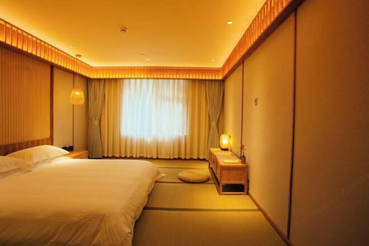 Une chambre d’hôtel avec un grand lit, une table de chevet avec une lampe, une fenêtre avec des rideaux et un sol traditionnel japonais recouvert de tatamis.