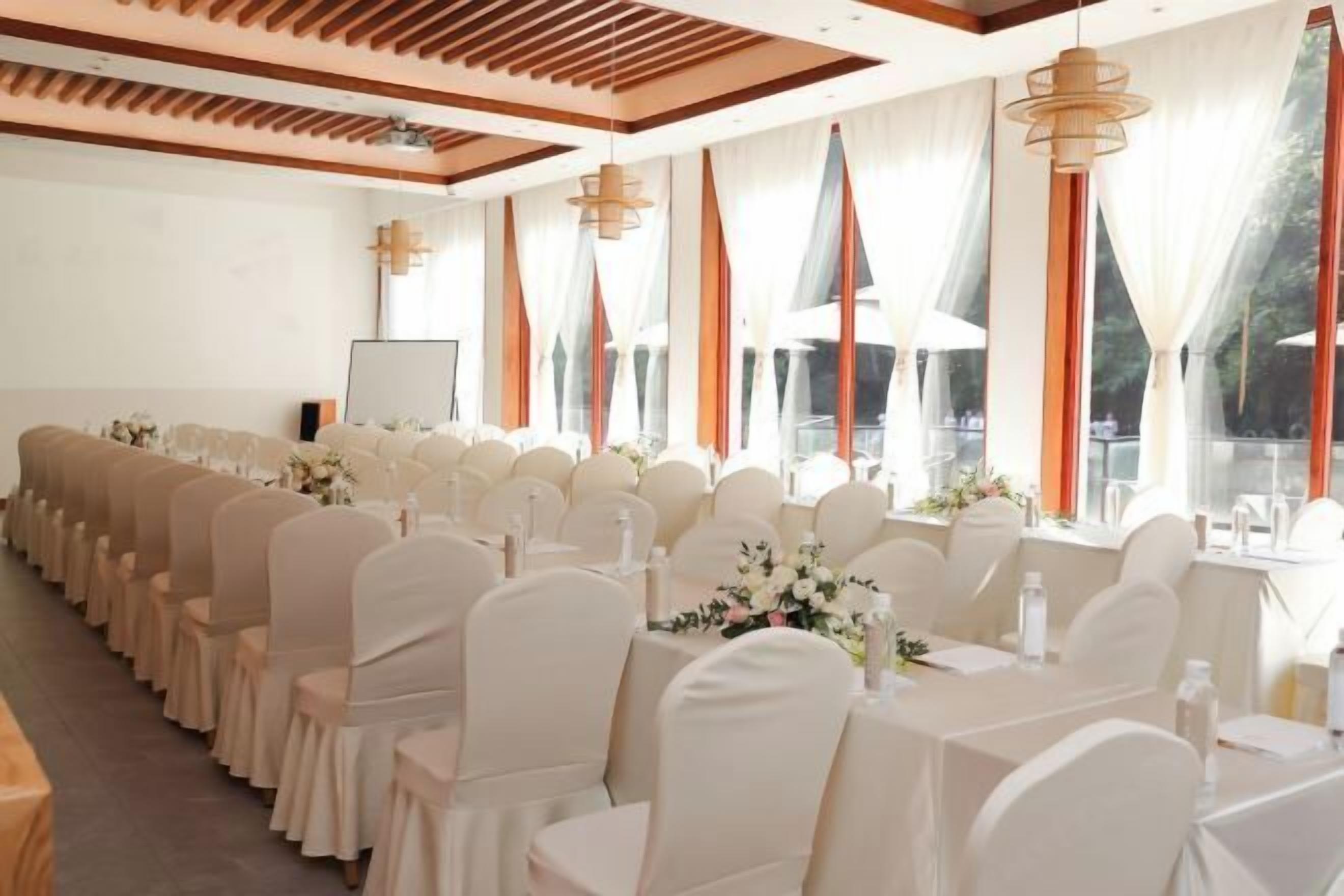 A banquet hall with rows of tables and chairs, large windows with sheer curtains, and pendant lighting.