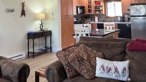 A spacious living area with a brown leather sofa, a wooden coffee table, and a patterned throw pillow. A dining table with white chairs is positioned near a kitchen area with wooden cabinets, a microwave, and a refrigerator. - Escape to the Pocono Mountains. (Tannersville)