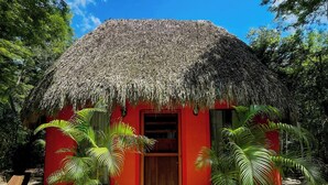 Una cabaña con techo de paja y exterior rojo, rodeada de palmeras y un sendero de grava.