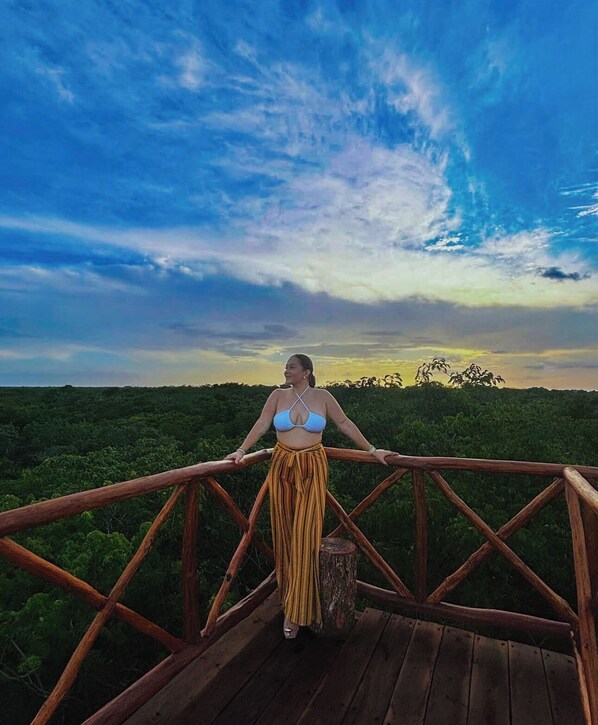 Una persona de pie en una terraza de madera con vista a un paisaje boscoso bajo un cielo parcialmente nublado.