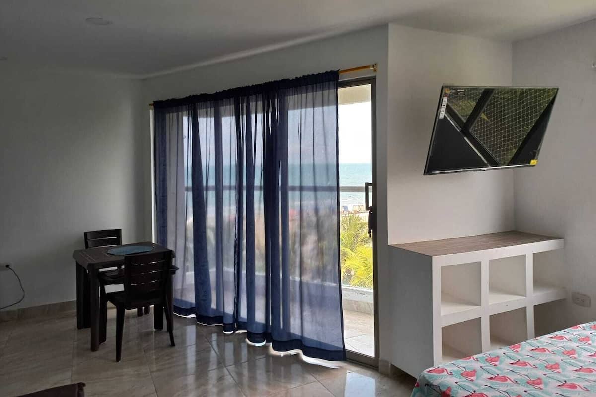 Une pièce comprenant une table à manger, des chaises, un lit recouvert d’une couvre-lit à motifs, un téléviseur fixé au mur et une vue sur la mer, visible à travers une fenêtre ornée de rideaux légers.