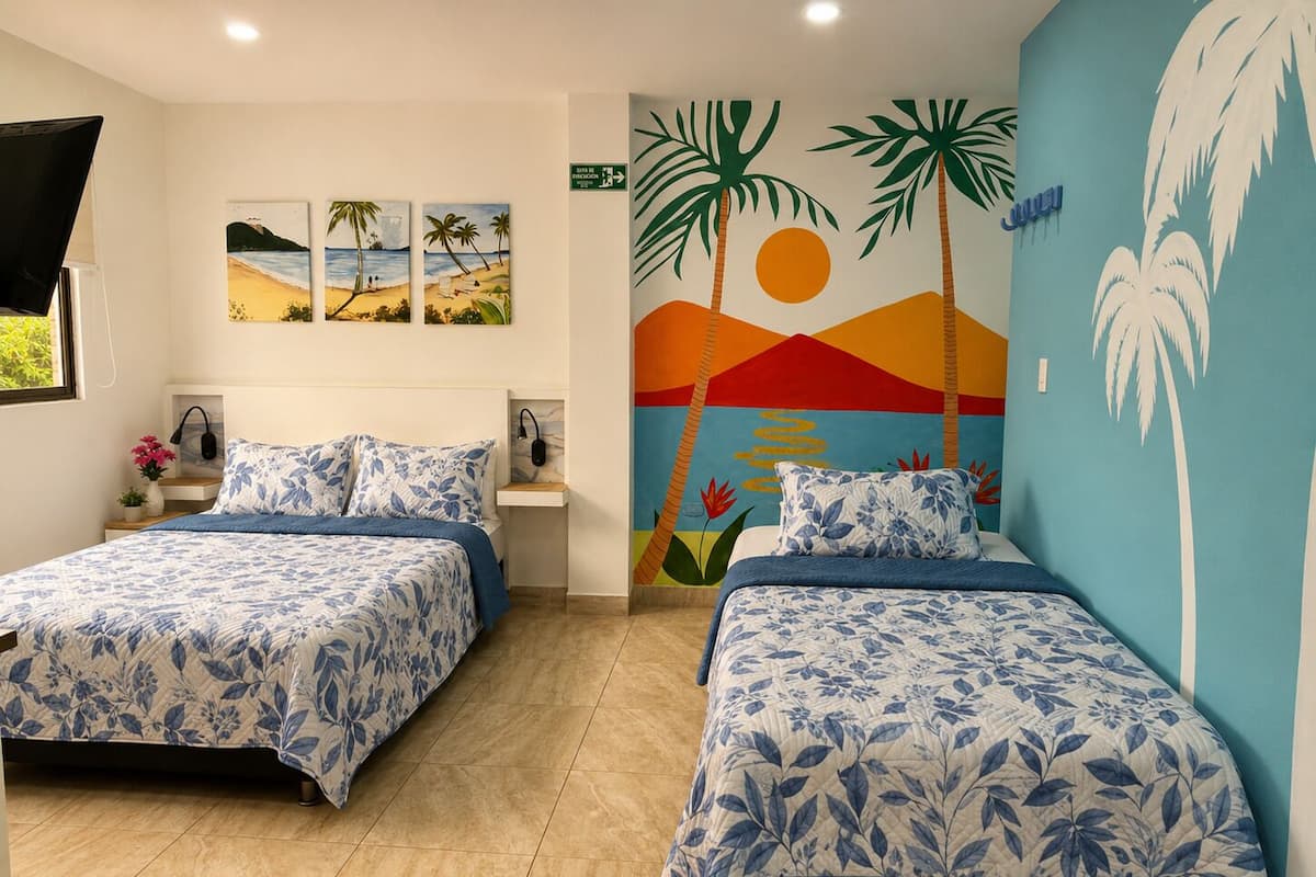 Une chambre d’hôtel avec deux lits, une fresque représentant des palmiers et un coucher de soleil, et des œuvres d’art accrochées au mur.