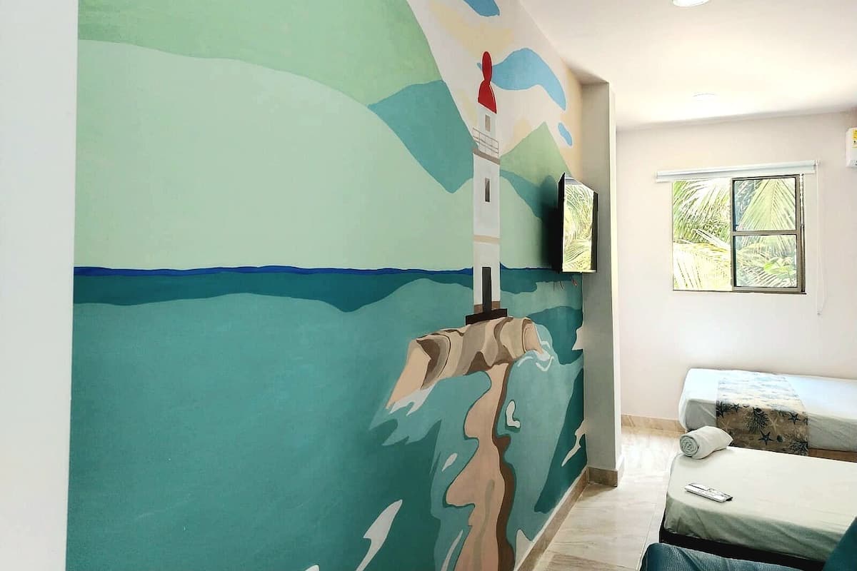 Une chambre décorée d’une fresque représentant un phare, avec un lit, un téléviseur fixé au mur et une fenêtre donnant sur des palmiers.