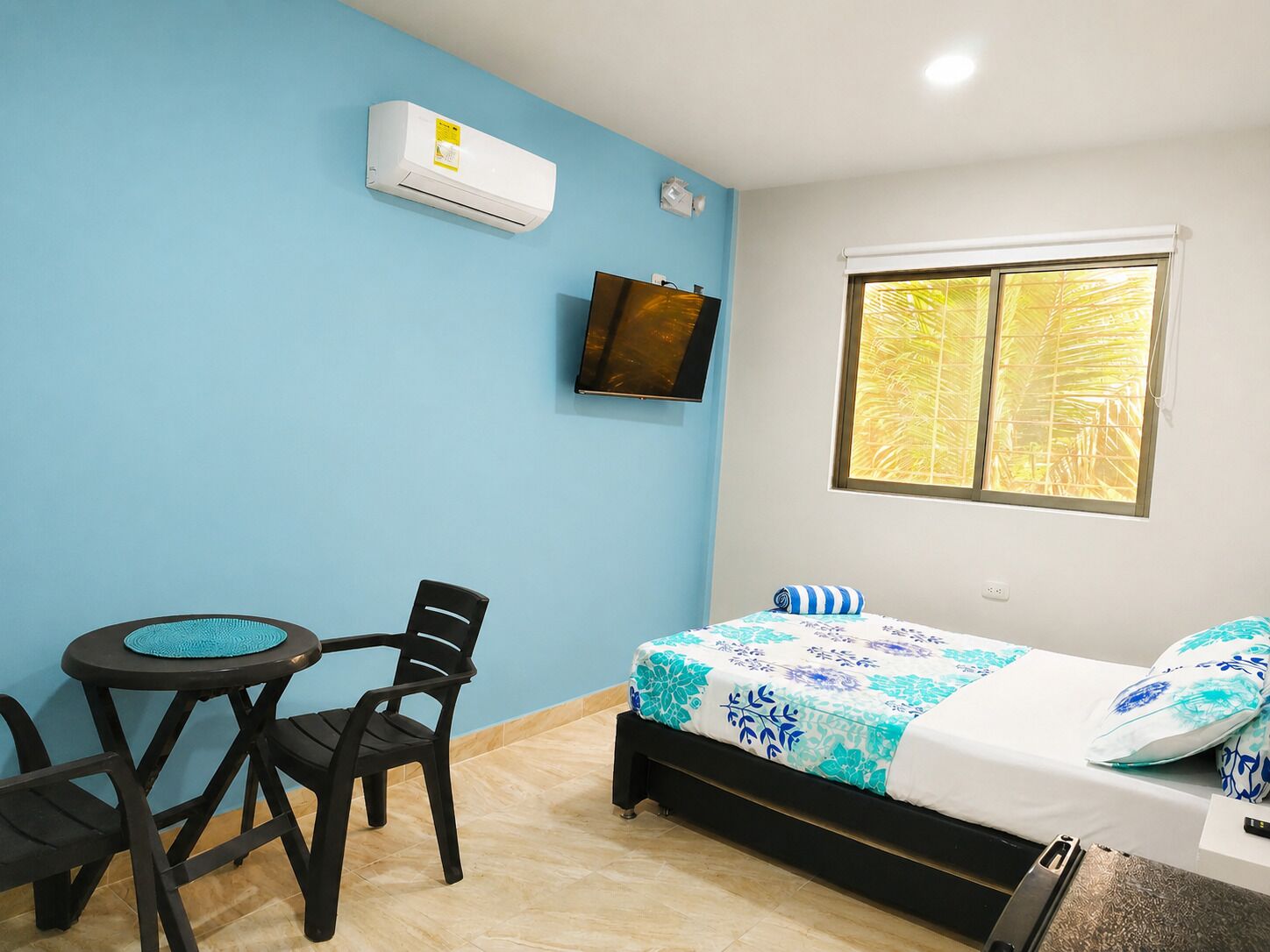 Una habitación con cama, una mesita con sillas, aire acondicionado, televisión y ventana con vistas a palmeras.
