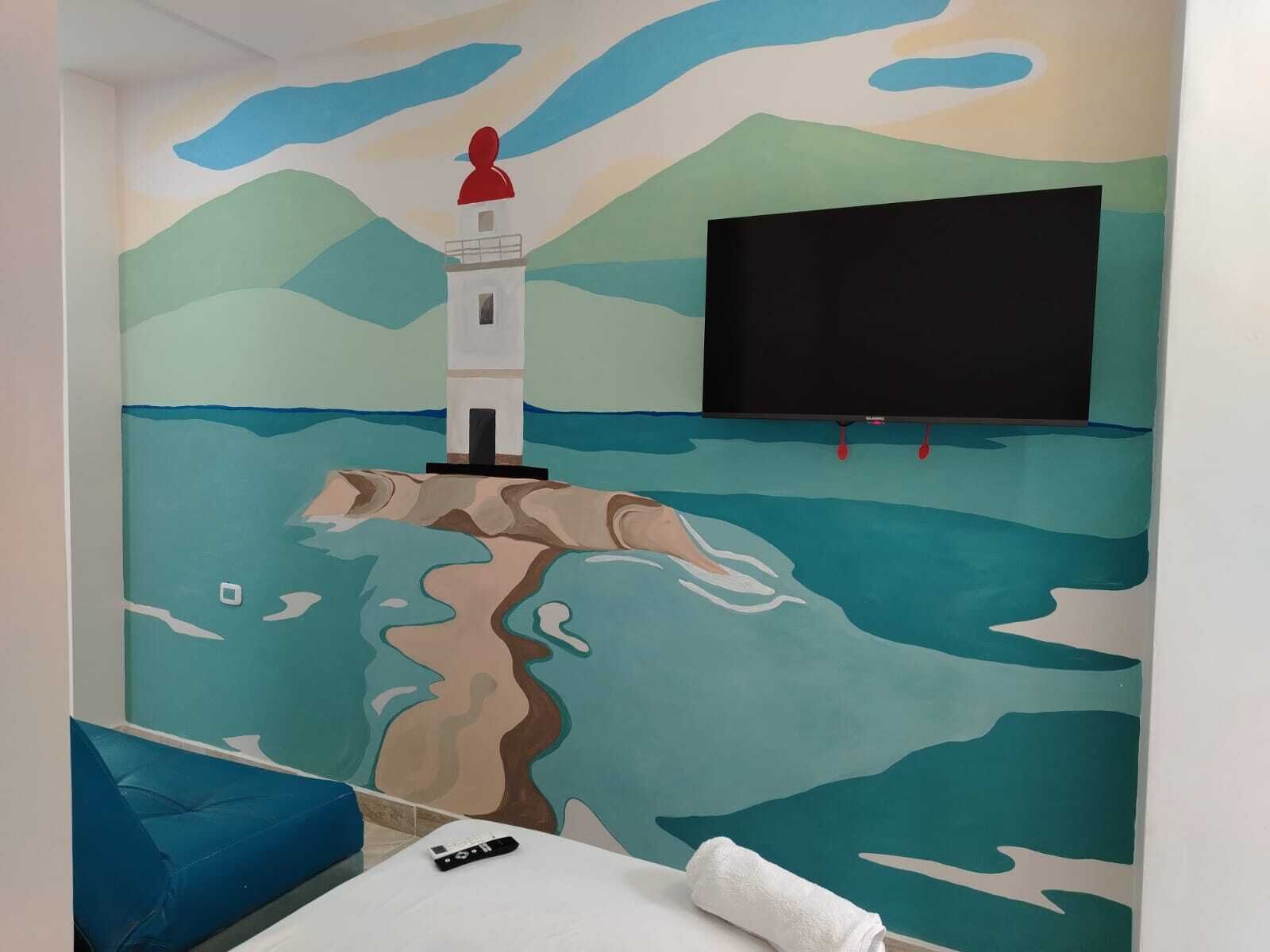Habitación con un mural de un faro sobre un acantilado, un televisor de pantalla plana en la pared y un sofá azul.