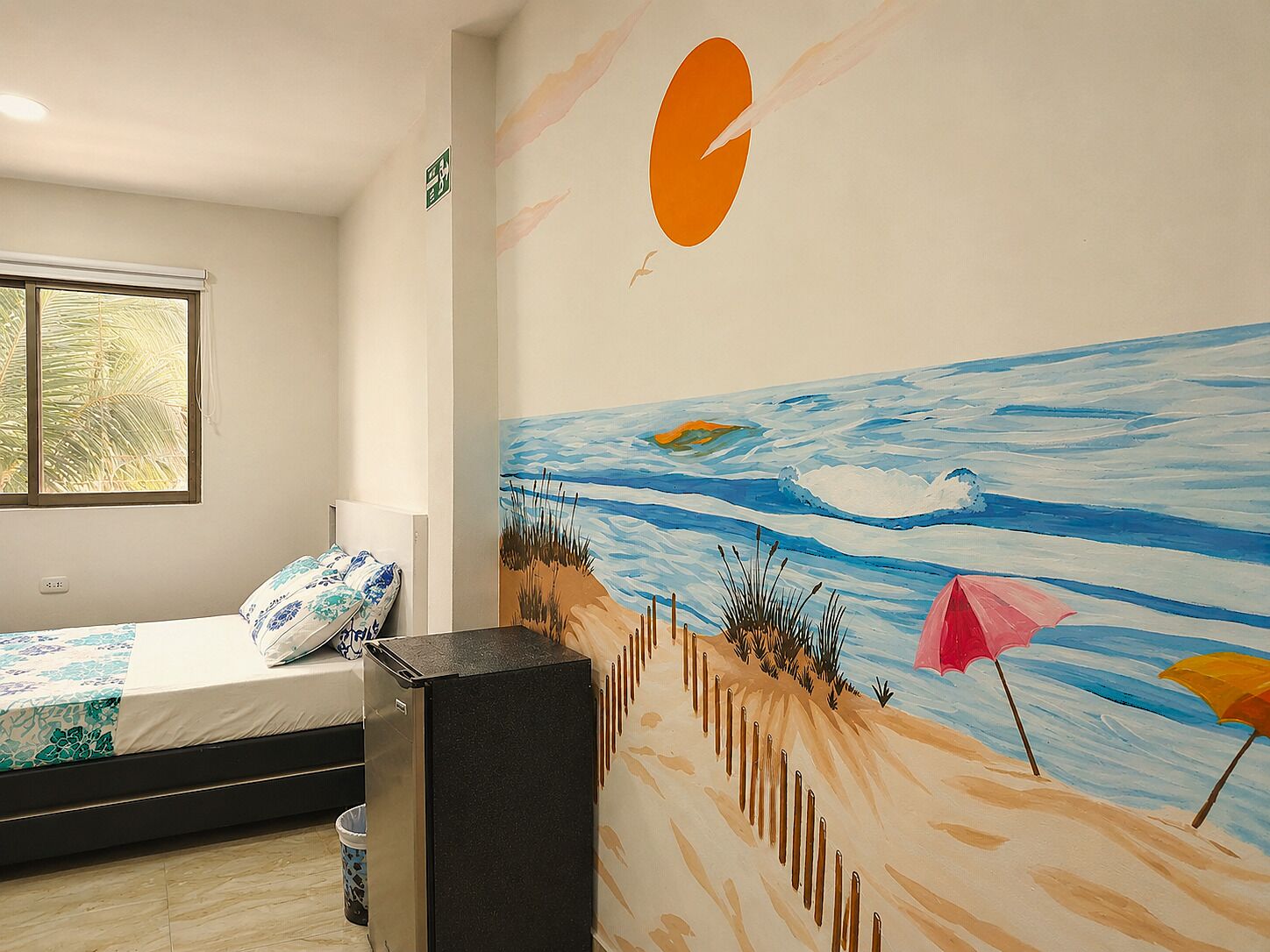 Un dormitori amb llit, finestra, mural a la paret i paperera.