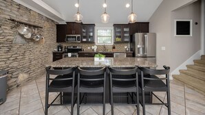 Una cocina con encimeras de granito, armarios oscuros, una pared de acento de piedra, una isla central con taburetes de bar e iluminación colgante.