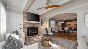 Una sala de estar con chimenea, ventilador de techo, comedor con mesa y sillas, y un televisor grande montado en la pared.