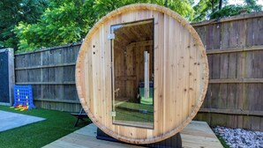 Una sauna de madera con puerta de vidrio, ubicada en un muelle de madera en un patio trasero.