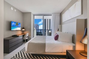 A modern hotel room with a large bed, a desk, a TV, and a balcony with a city view.