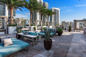 A rooftop lounge with outdoor seating, palm trees, and a cityscape view.