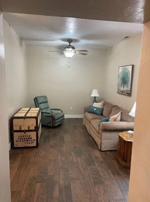Cozy living space with a sofa, armchair, and wooden side table, featuring a ceiling fan for ventilation and a decorative painting.