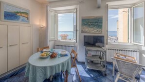 A dining area with a table set for two, a television, and a view of the sea through open windows.