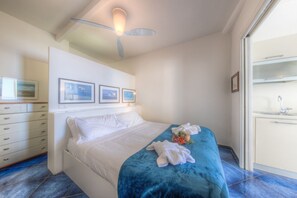 Ein Schlafzimmer mit Bett, Schminktisch, Deckenventilator, gerahmten Bildern an der Wand sowie einem Badezimmer mit Waschbecken und Spiegel.
