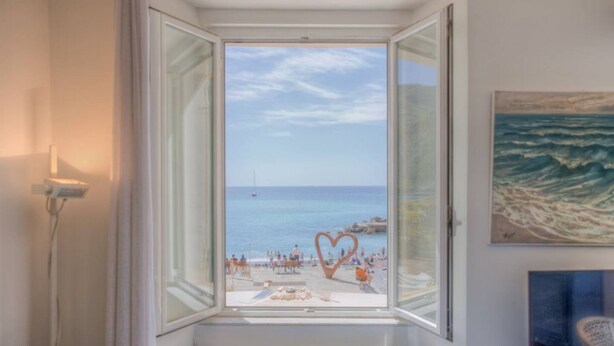 A room with a bay window overlooking a beach, a radiator below, and a painting on the wall.