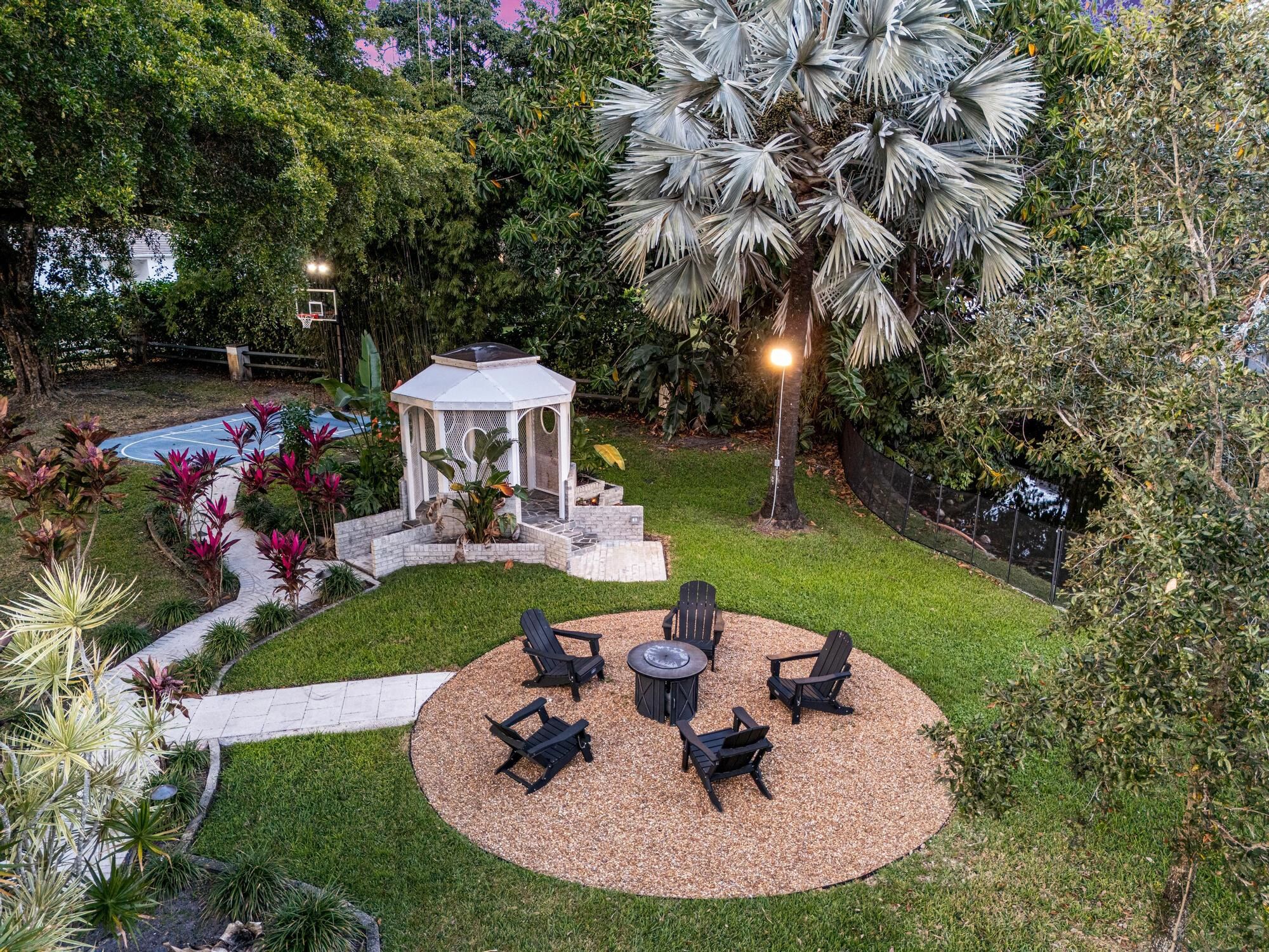 A circular outdoor seating area with a fire pit and four chairs, surrounded by a gravel path and lush greenery.