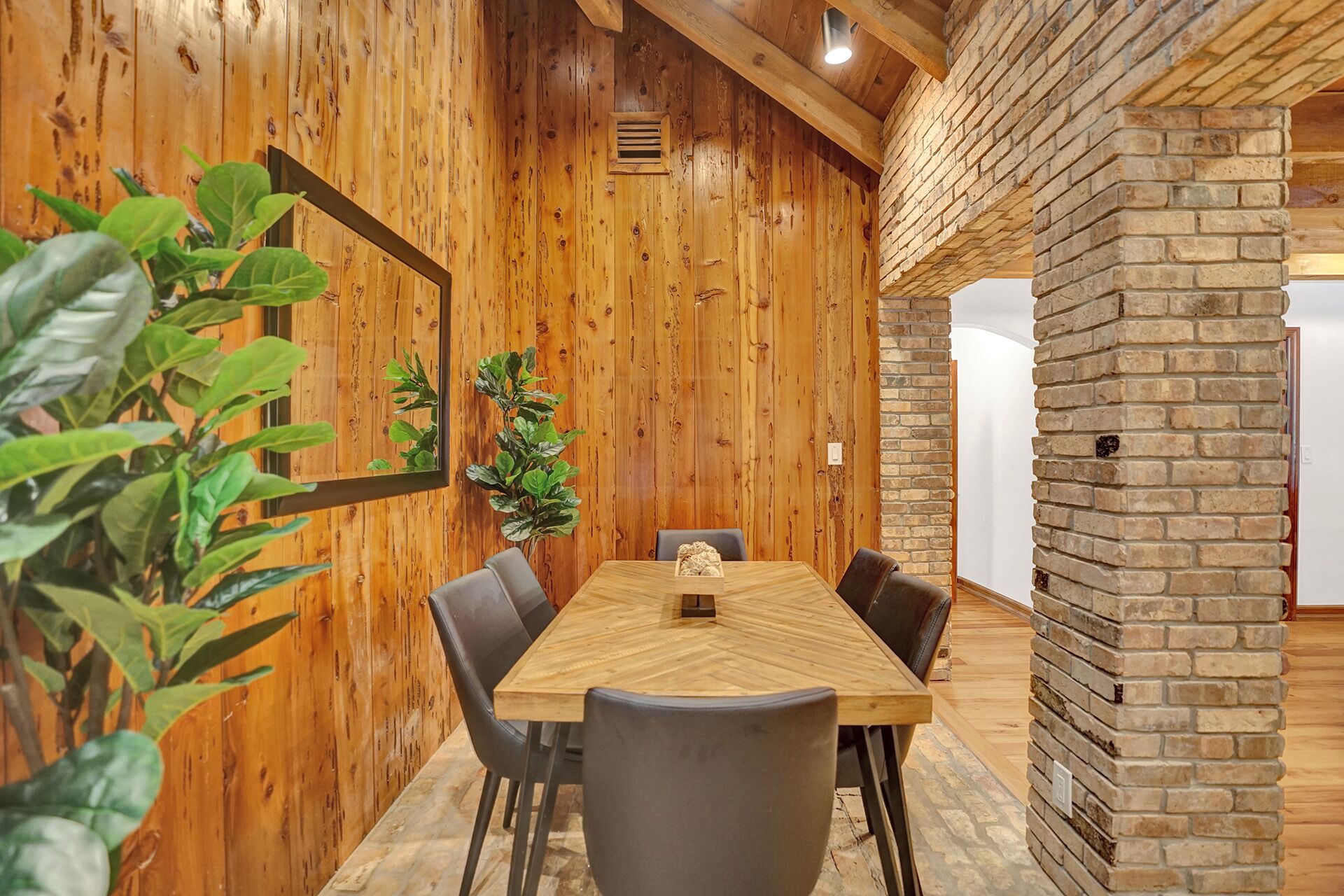 A wooden-paneled room with a large table and chairs, a potted plant, and a brick wall.