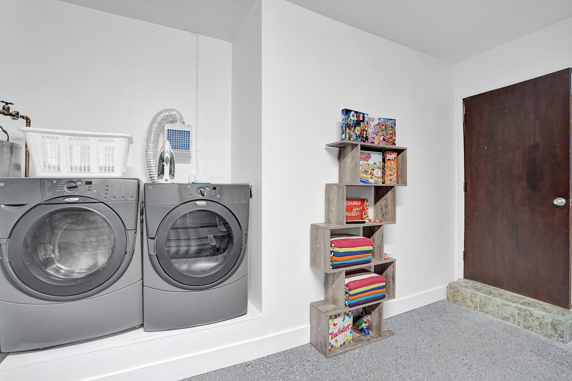 A laundry area with two front-loading washing machines, a drying rack, and a wooden shelf unit with folded towels and children's board games.