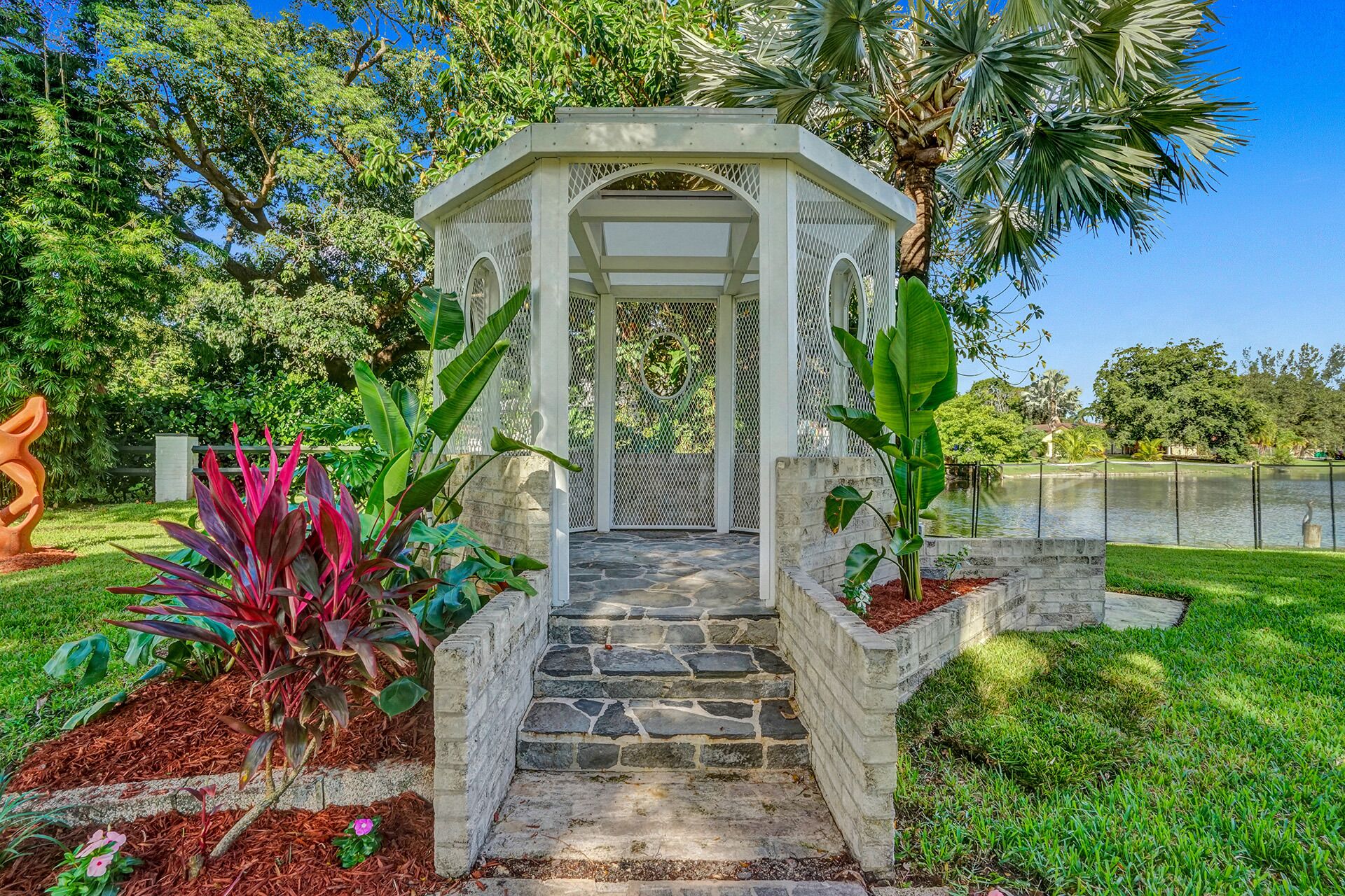 A gazebo with stone steps leading up to it, surrounded by lush greenery and a body of water in the background.