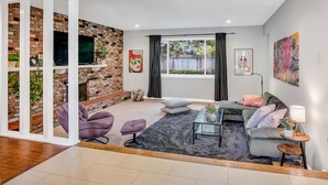 A modern living room with a brick wall, a flat-screen TV, a grey sofa, a coffee table, and a window with curtains. - Large home - Sleeps many 4B/2B (Walnut Creek)