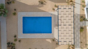 Eine Vogelperspektive auf einen rechteckigen Pool, umgeben von Sand, mit einem schattigen Sitzbereich auf der rechten Seite.