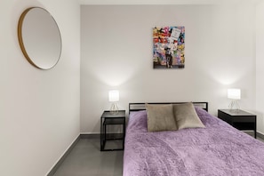 Ein Schlafzimmer mit einem Bett, zwei Nachttischen, einem Wandspiegel und einem farbenfrohen Kunstwerk.