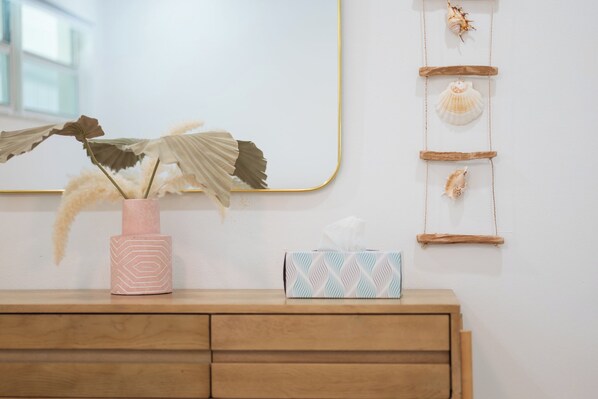 A wooden dresser with a pink vase containing dried plants, a tissue box, and a wall-mounted shelf with seashells. - Jetties Pointe Cottage #4 - 408 “Casey Key” (Nokomis)