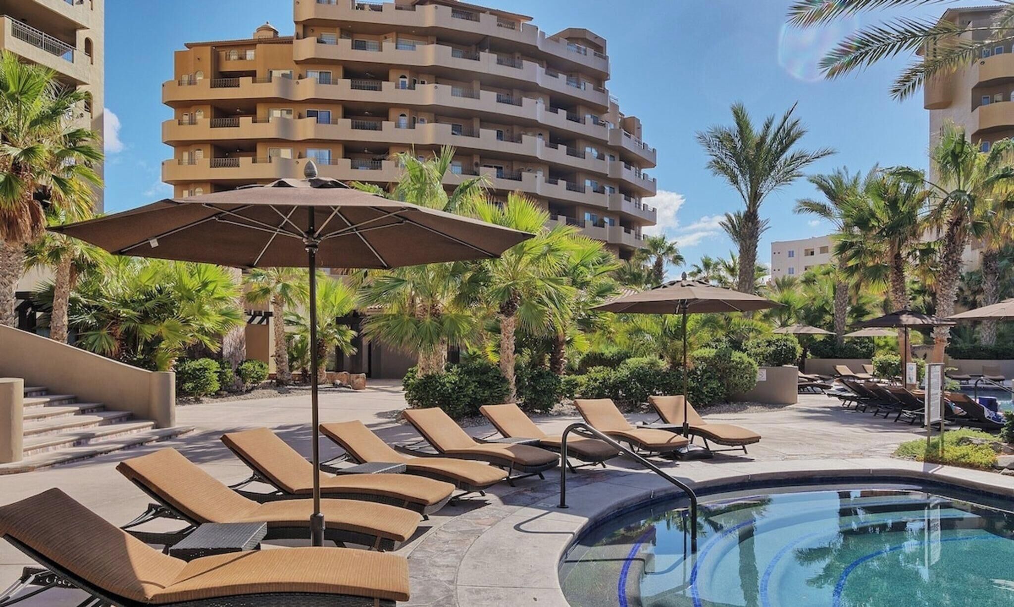 Ein Hotelpoolbereich mit Liegestühlen und Sonnenschirmen, umgeben von Palmen und einem mehrstöckigen Gebäude.