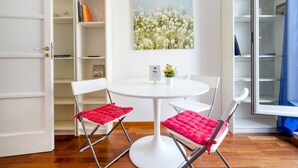 Habitació amb taula rodona blanca, dues cadires amb coixinets vermells, una prestatgeria i un quadre amb flors.