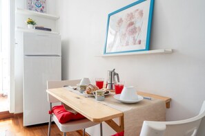 Une petite cuisine équipée d’un réfrigérateur blanc, d’une table en bois recouverte d’une nappe beige, et d’un coin petit-déjeuner comprenant des croissants, des tasses à café et un pichet à jus.