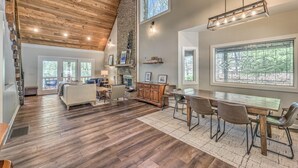 A spacious living area with a high ceiling, wooden flooring, a dining table with chairs, and large windows offering views of the outdoors. - Lake Cove Oasis - Waterfront on Lake Blue Ridge | Private Yard and Dock | Pet Friendly (Morganton)