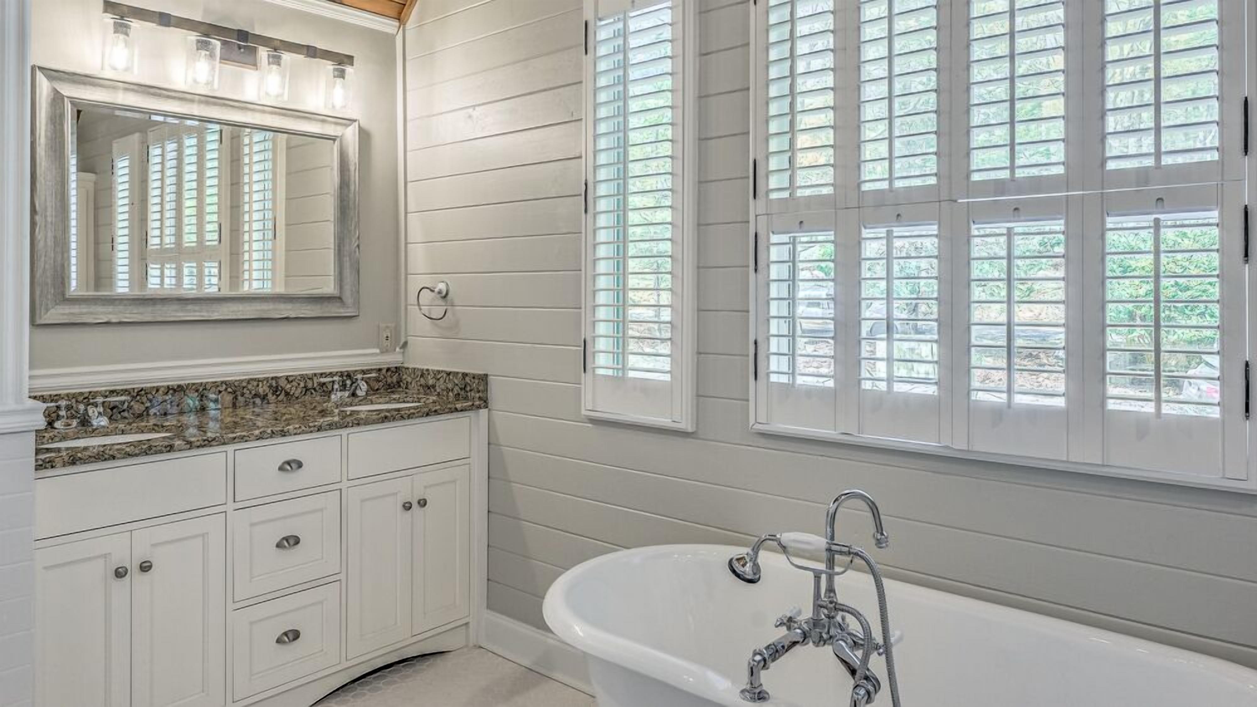 A bathroom with a freestanding bathtub, a vanity with drawers, a large mirror, and a window with shutters.