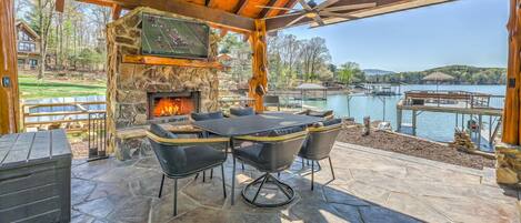 Una chimenea de piedra con un televisor montado encima, rodeada de asientos exteriores con vista a un lago y montañas.