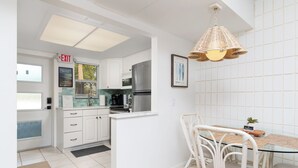 Een moderne keuken met witte kasten, een roestvrijstalen koelkast, en een eethoek met een glazen tafel en rieten stoelen.