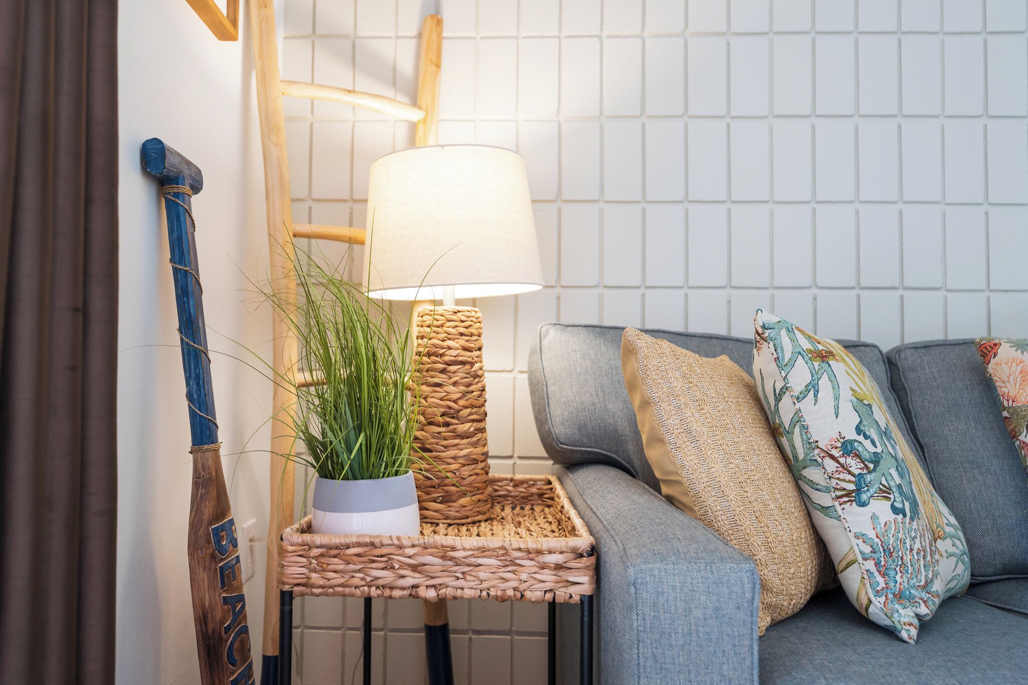 Ein Zimmer mit grauem Sofa, einem gewebten Beistelltisch mit Lampe und einer Topfpflanze.