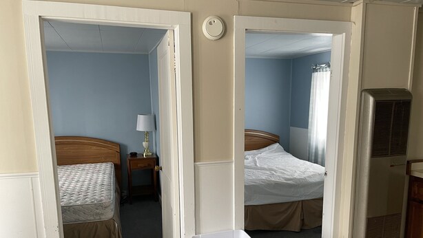 A hotel room with two beds, a bedside table with a lamp, a radiator, and a trash bin. - Autrain Motel (Au Train)