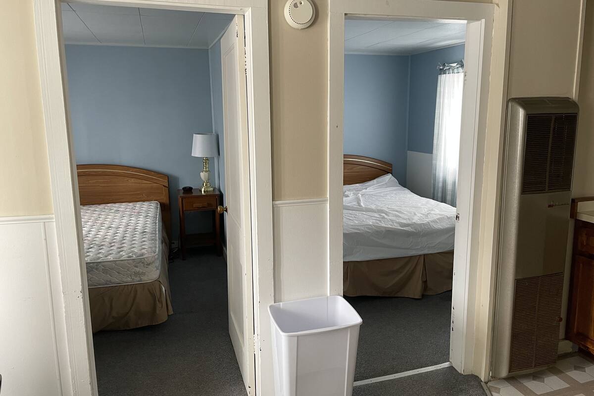 Quarto de hotel com duas camas, uma mesa de cabeceira com luminária, um radiador e uma lixeira.