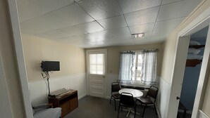 A small, compact room with a single bed, a desk with two chairs, a television mounted on the wall, and a window with curtains. - Autrain Motel (Au Train)