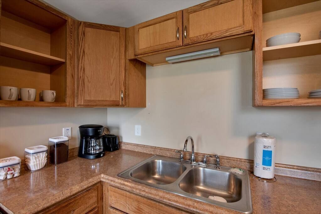 A kitchen with wooden cabinets, a double sink, a coffee maker, and a shelf with dishes.