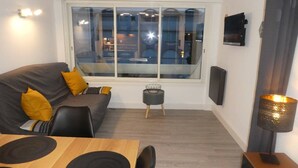 Ein modernes Wohnzimmer mit einem grauen Sofa, gelben Kissen, einem kleinen runden Tisch und einem an der Wand befestigten Fernseher.