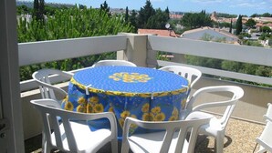 Ein runder Tisch mit blauer Tischdecke, verziert mit Sonnenblumen, umgeben von weißen Plastikstühlen auf einem Balkon mit Blick auf Häuser und Grünflächen.