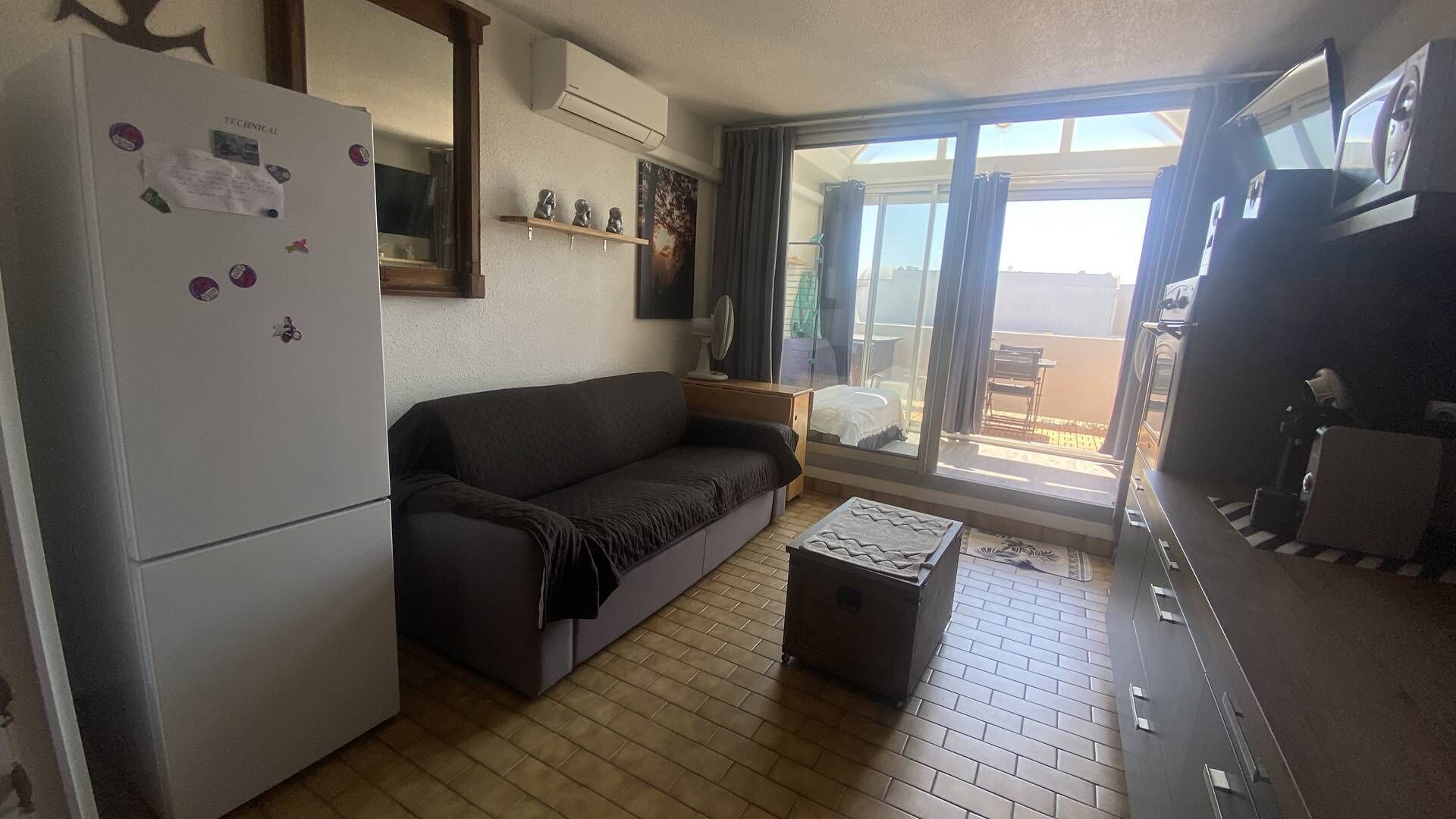 Un espace de vie compact comprenant un réfrigérateur, un canapé, une petite table et une porte-fenêtre coulissante donnant sur un espace extérieur.