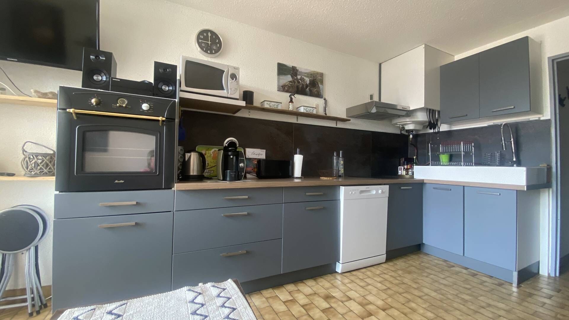 A compact kitchen with a built-in oven, microwave, and dishwasher. There is a clock on the wall and a small shelf with decorative items.