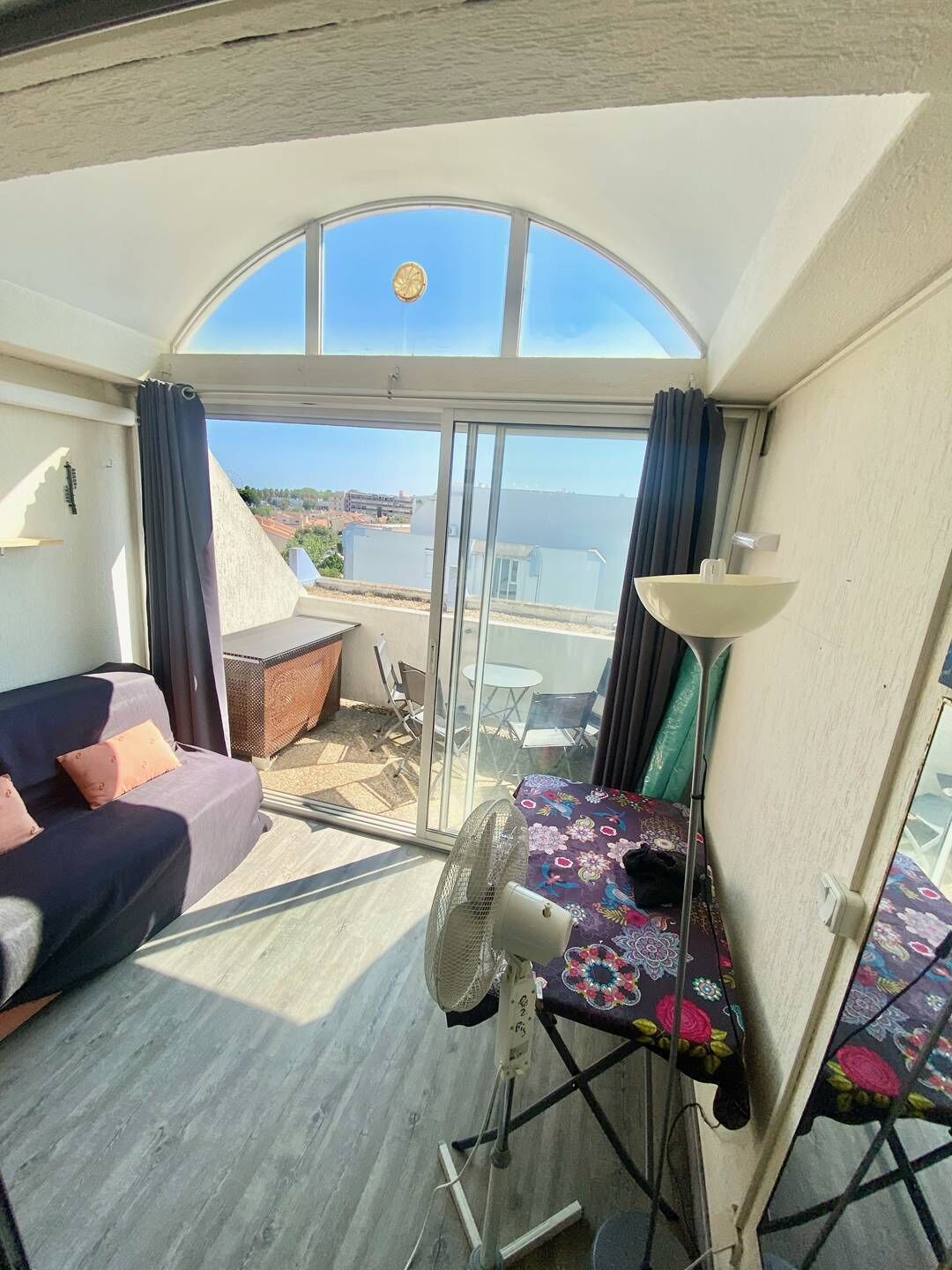 A balcony with a sofa, a table with a tablecloth, a fan, and a view of the sea.