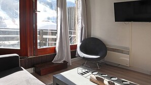 Ein Zimmer mit großem Fenster, einem Flachbildfernseher, einem modernen Sessel und einem kleinen Tisch mit Kerzenhalter.