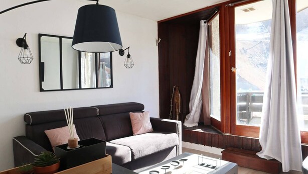 Ein modernes Wohnzimmer mit einem dunklen Sofa, einem Couchtisch mit Weingläsern und einem großen Fenster mit Vorhängen.